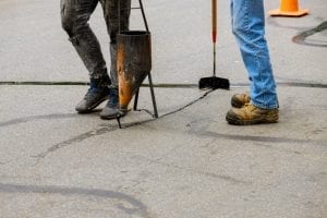 two workers in work boots sealing cracks in asphalt pavement with hot tar