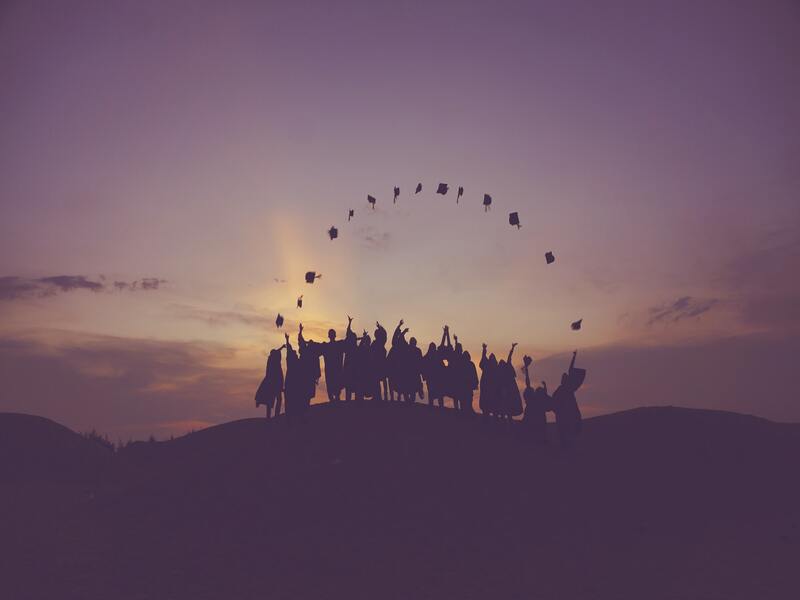 A large group of people on a hillside at sunset celebrating by throwing hats into the air