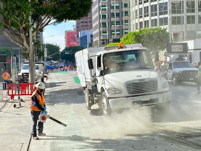 A sweeper truck sweeping a city street while another worker in a safety vest and hard hat operates a blower to remove litter and debris