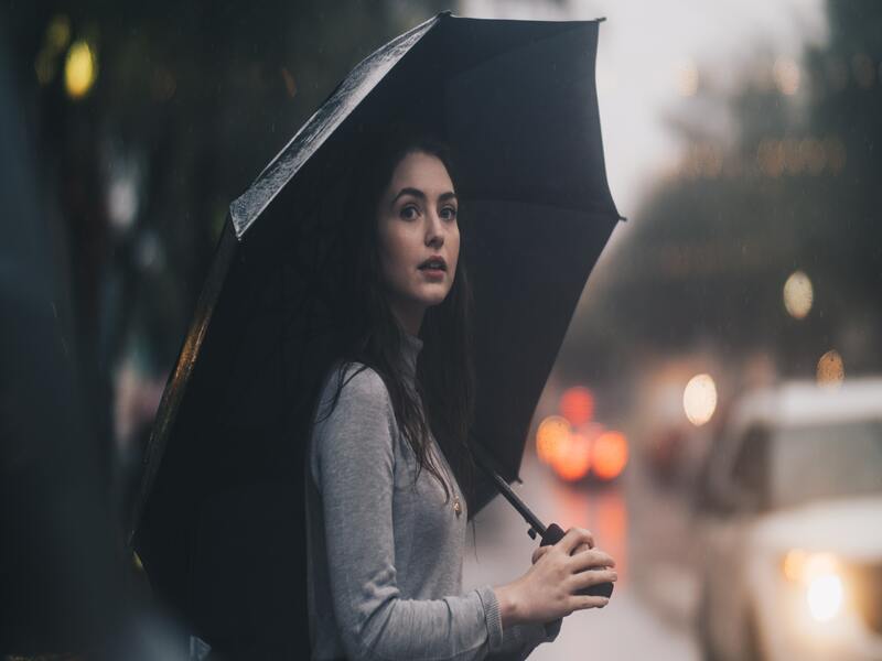 Yoiung woman with dark hair walking a city street in the rain while carrying an umbrella