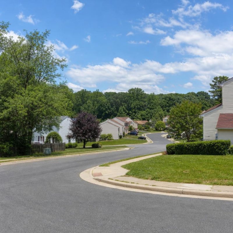Rolling, wooded suburban street in Dale City with curving pavement, detached homes, and townhouse clusters.